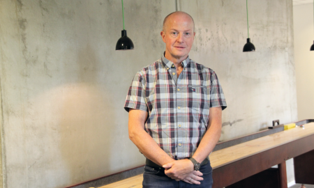 En mann står i et rom med betongvegg, iført skjorte med rutemønster og har på seg et armbånd, foran en lang benk og med to svarte hengelamper over seg.