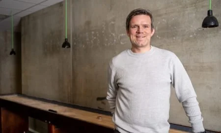 En mann står foran en bar eller bardisk i et rom med betongvegg. Han smiler og ser mot kamera.