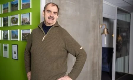 En mann står foran en grønn vegg med bilder og en skjerm, i en moderne innendørs setting.