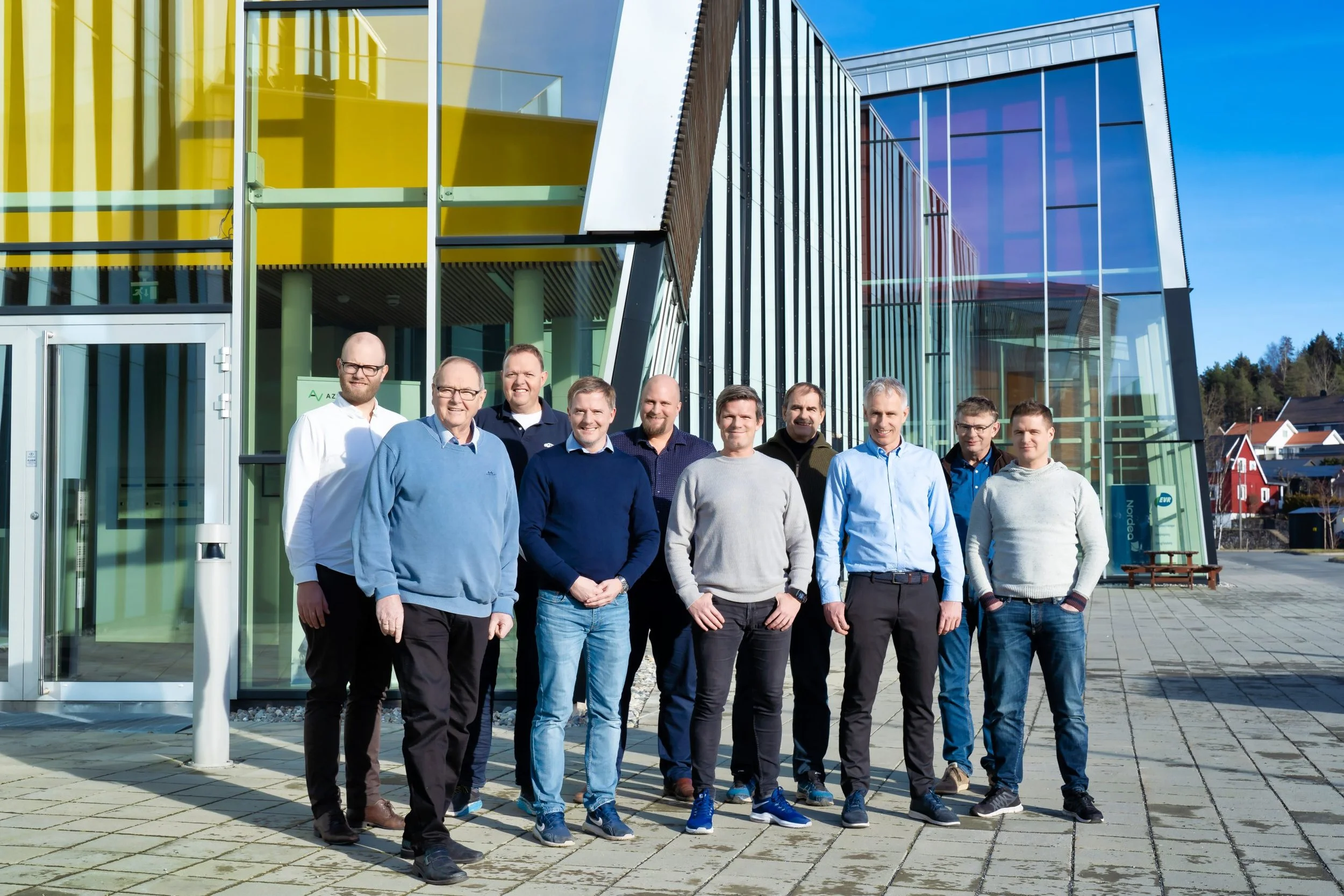 En gruppe menn står foran en moderne glassbygning på en solrik dag.