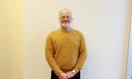 En mann med grått skjegg og briller, iført en gul genser, står foran en lys vegg og smiler.