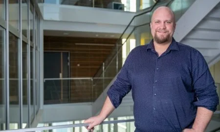 En mann med skjegg og blå skjorte står innendørs i et moderne kontormiljø.