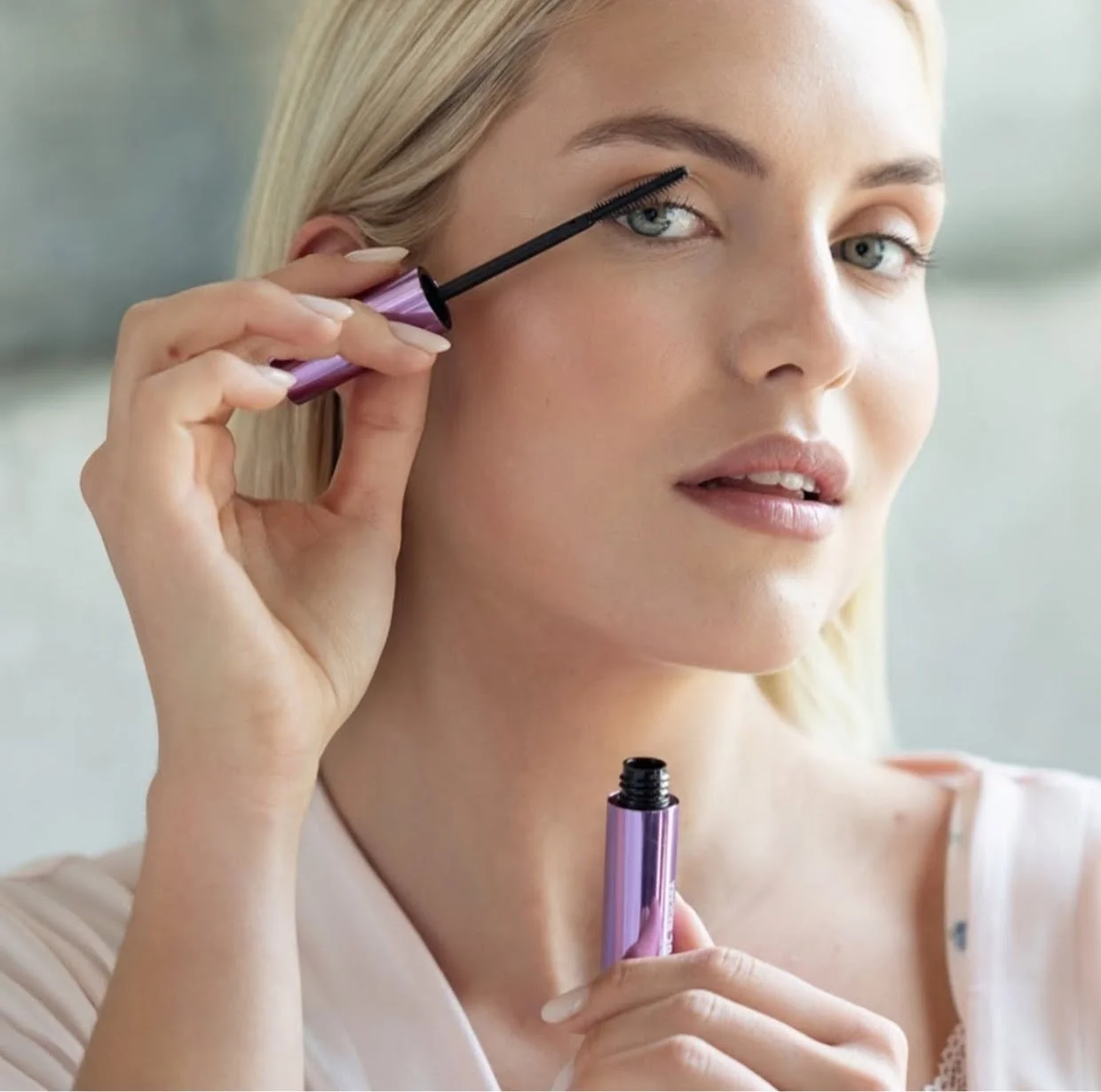 Een vrouw past mascara toe op haar wimperlijn met een mascara borstel, met de fles mascara in haar hand.