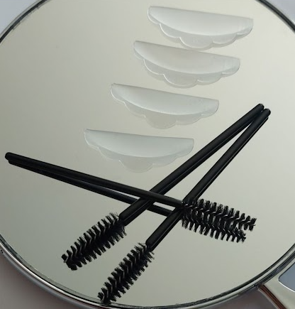 Twee zwarte mascara borstels en zes witte ellipsvormige oogmaskers op een spiegel.