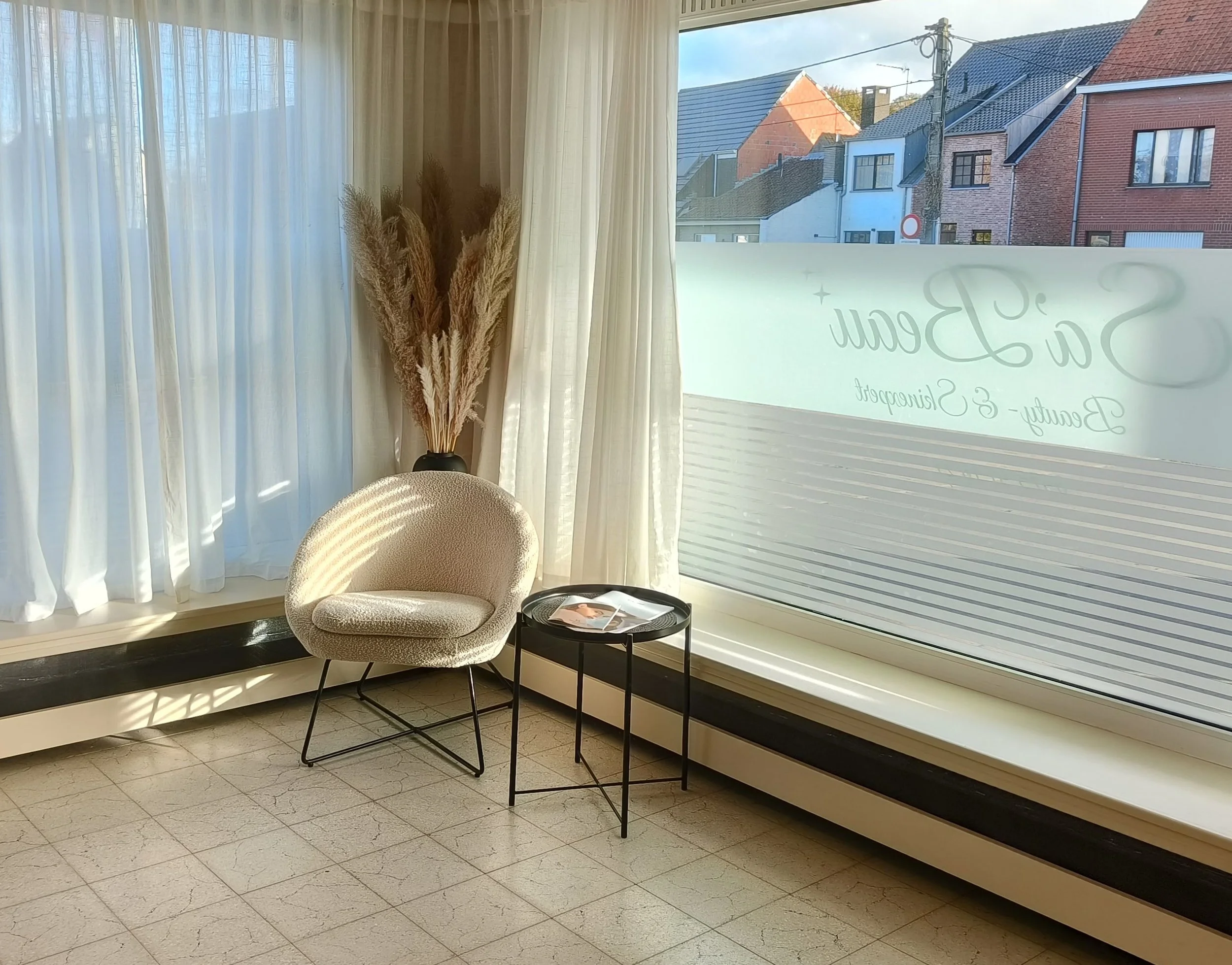 Een zittende stoel met een ronde rugleuning en een kussen, naast een kleine ronde tafel met tijdschriften, gelegen voor grote ramen met witte gordijnen en een grote vaas met gedroogde pampasgras binnenin. Buiten zijn huizen en een straat zichtbaar.