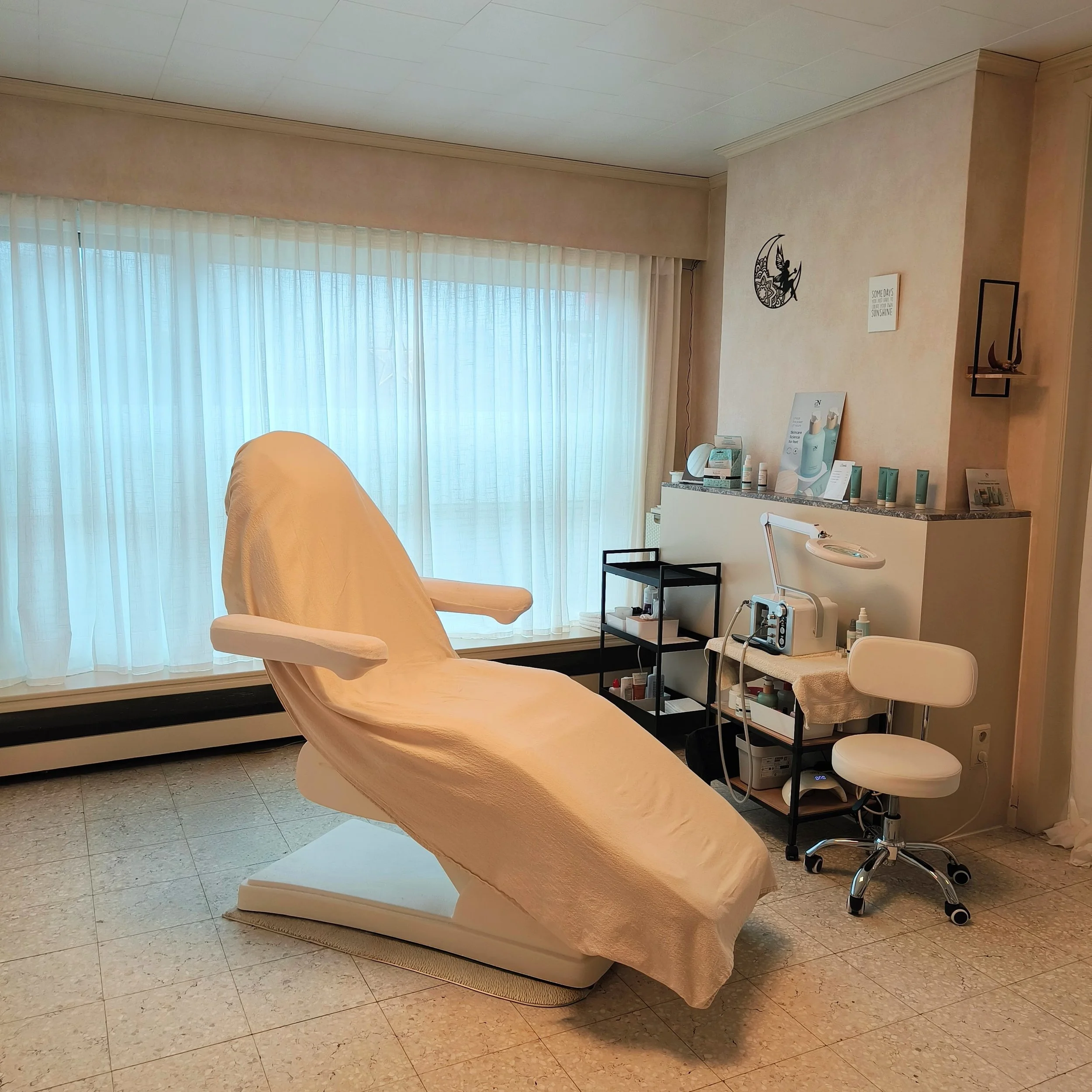 Kamer met een lege behandelingsbank en medische apparatuur, gekleed in beige met witte accessoires, binnen in een kliniek of salon.
