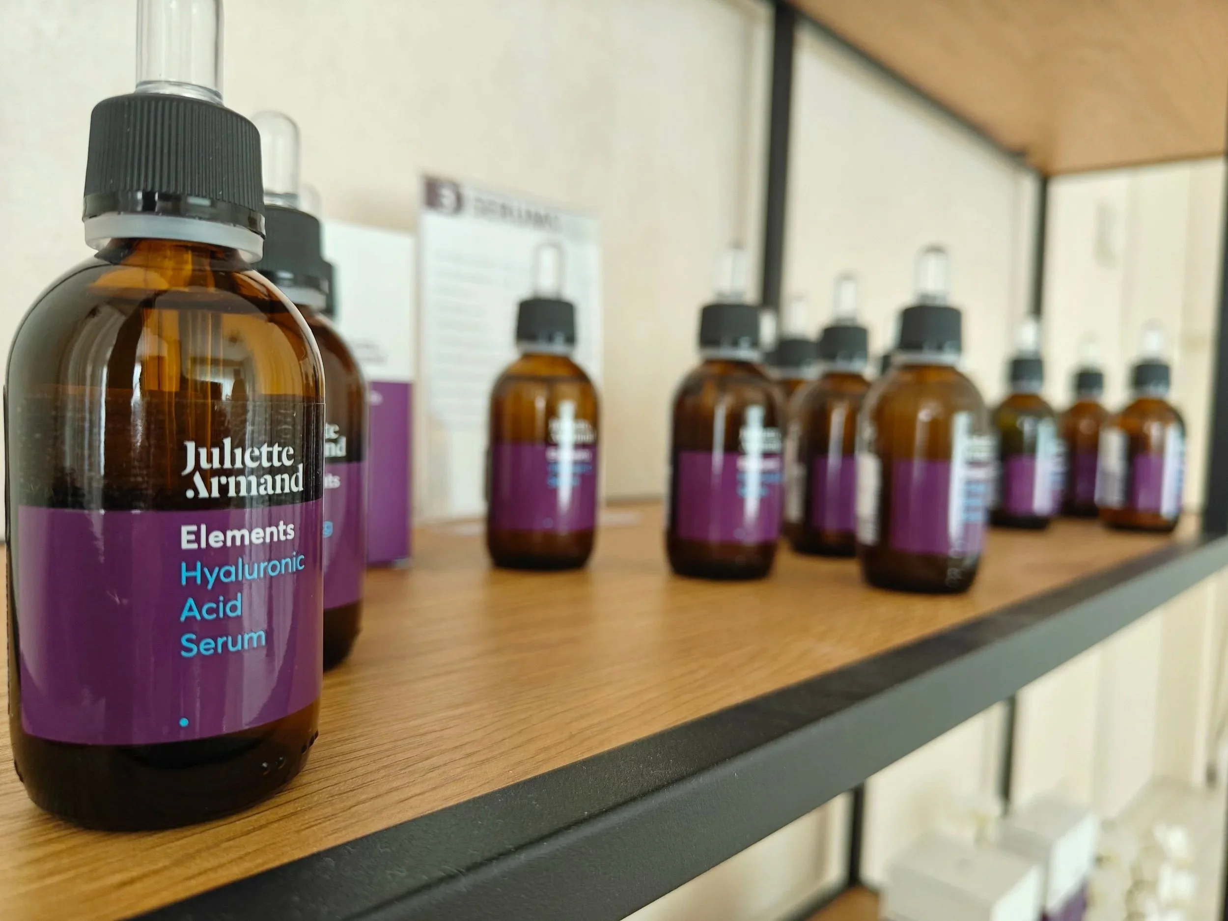 Reeks van Amberkleurige flesjes met zwarte doppen op een houten plank, het eerste flesje is duidelijk zichtbaar en heeft een label met de tekst 'Juliette Armand Elements Hyaluronic Acid Serum'.