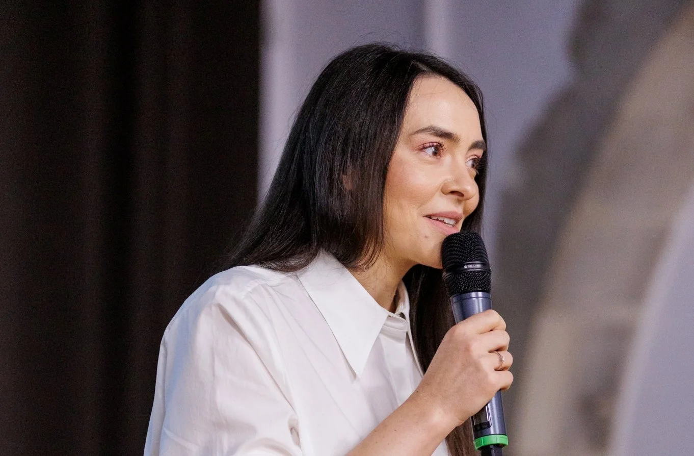 Woman with long dark hair holding a microphone and speaking at an indoor event.