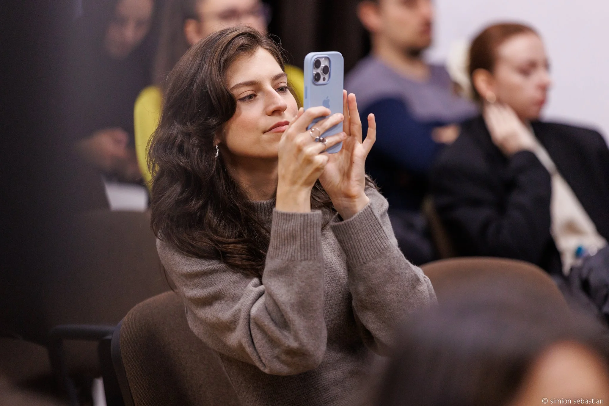 A woman with dark wavy hair sitting in a meeting or conference, holding and looking at her phone.