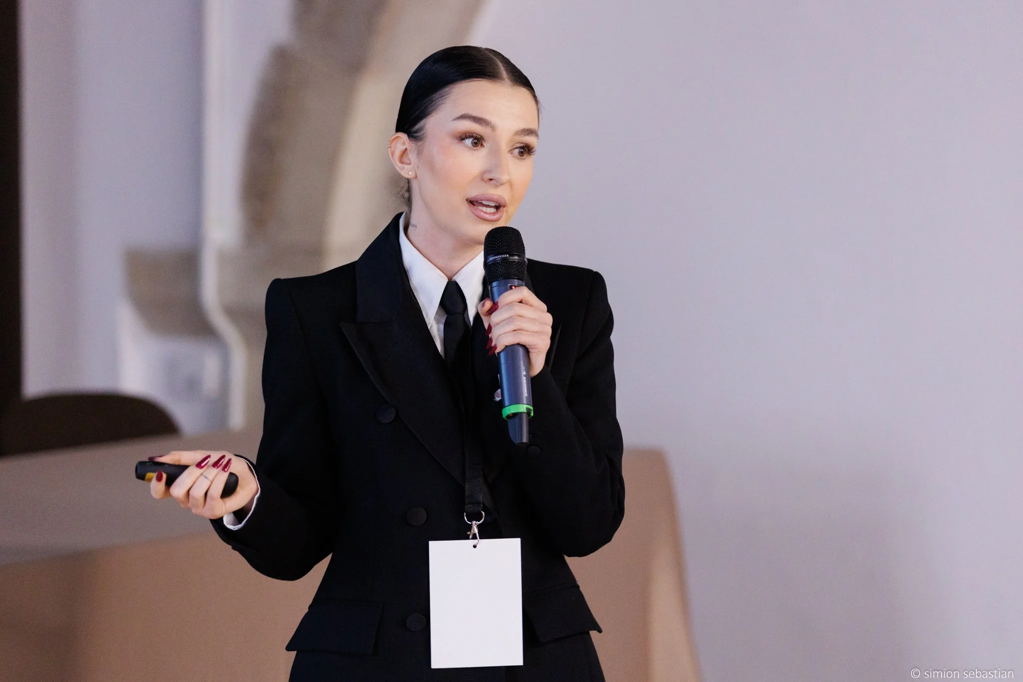 A woman in a black suit holding a microphone and a remote control, speaking in a conference or presentation setting.