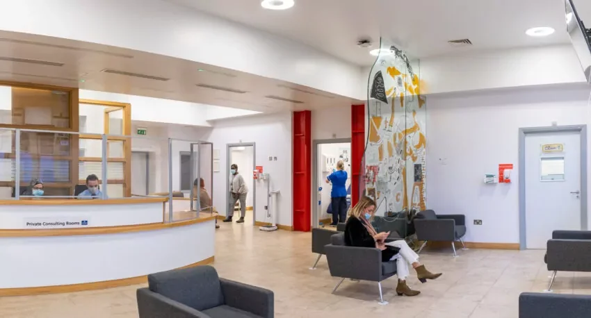 Hospital waiting area with people, some wearing masks, seated and standing near private consulting rooms, and a staff member at the reception desk.