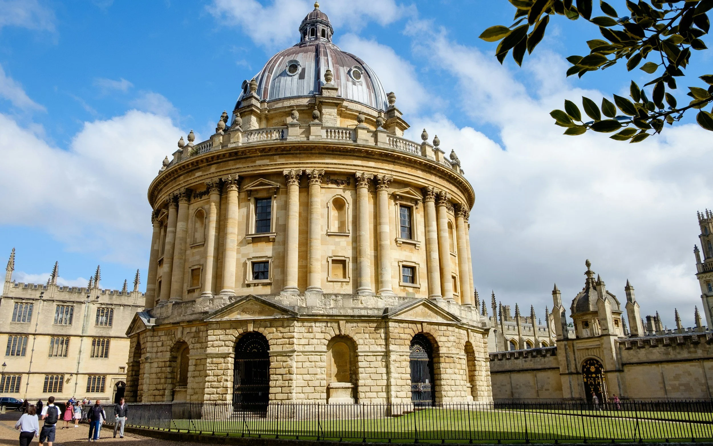 The dome of the Radcliffe Camera, a historic building in Oxford, surrounded by a fence with people walking nearby under a partly cloudy sky.