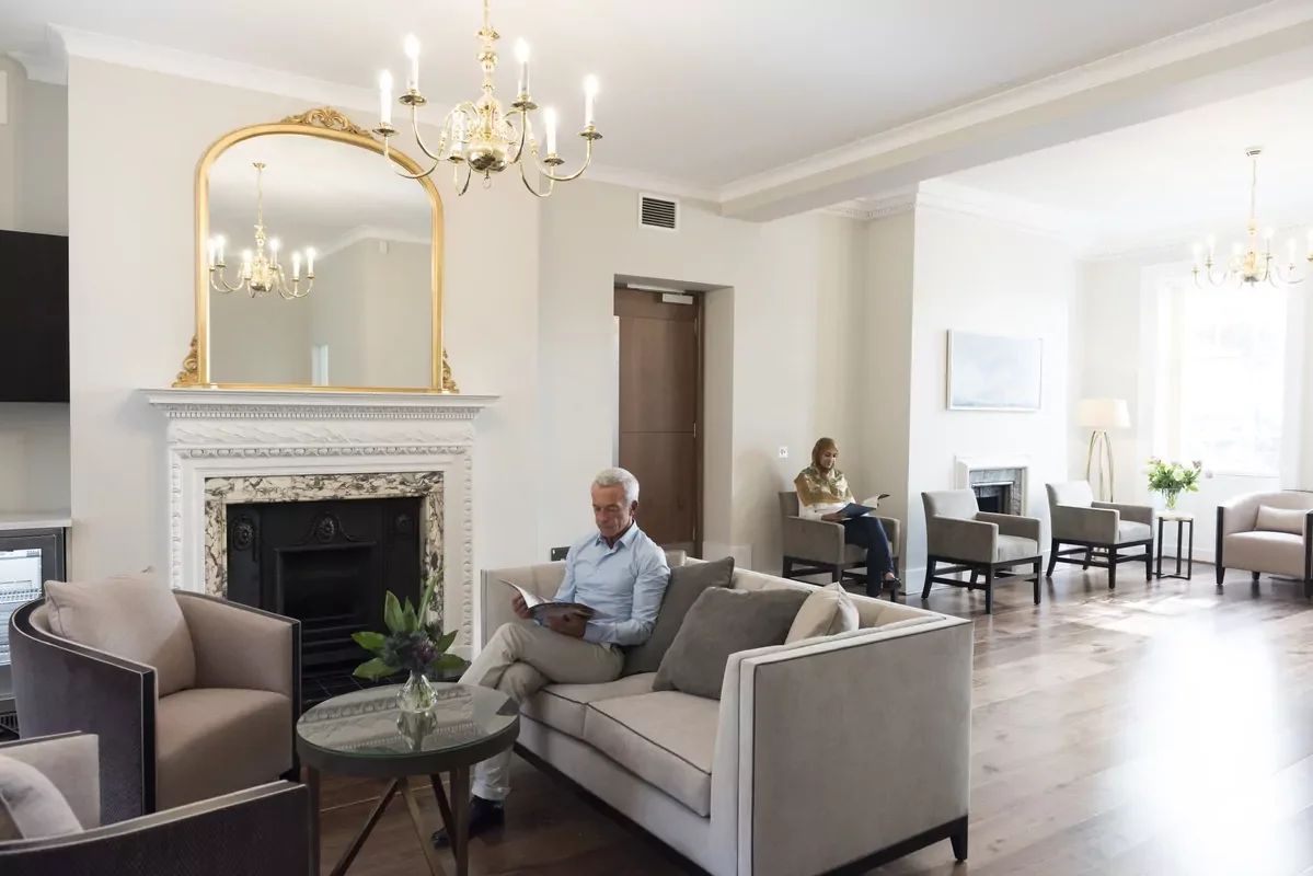 A bright, elegant waiting area with multiple chairs and sofas, a fireplace with a large mirror above, chandelier lighting, and three people seated reading magazines or books.