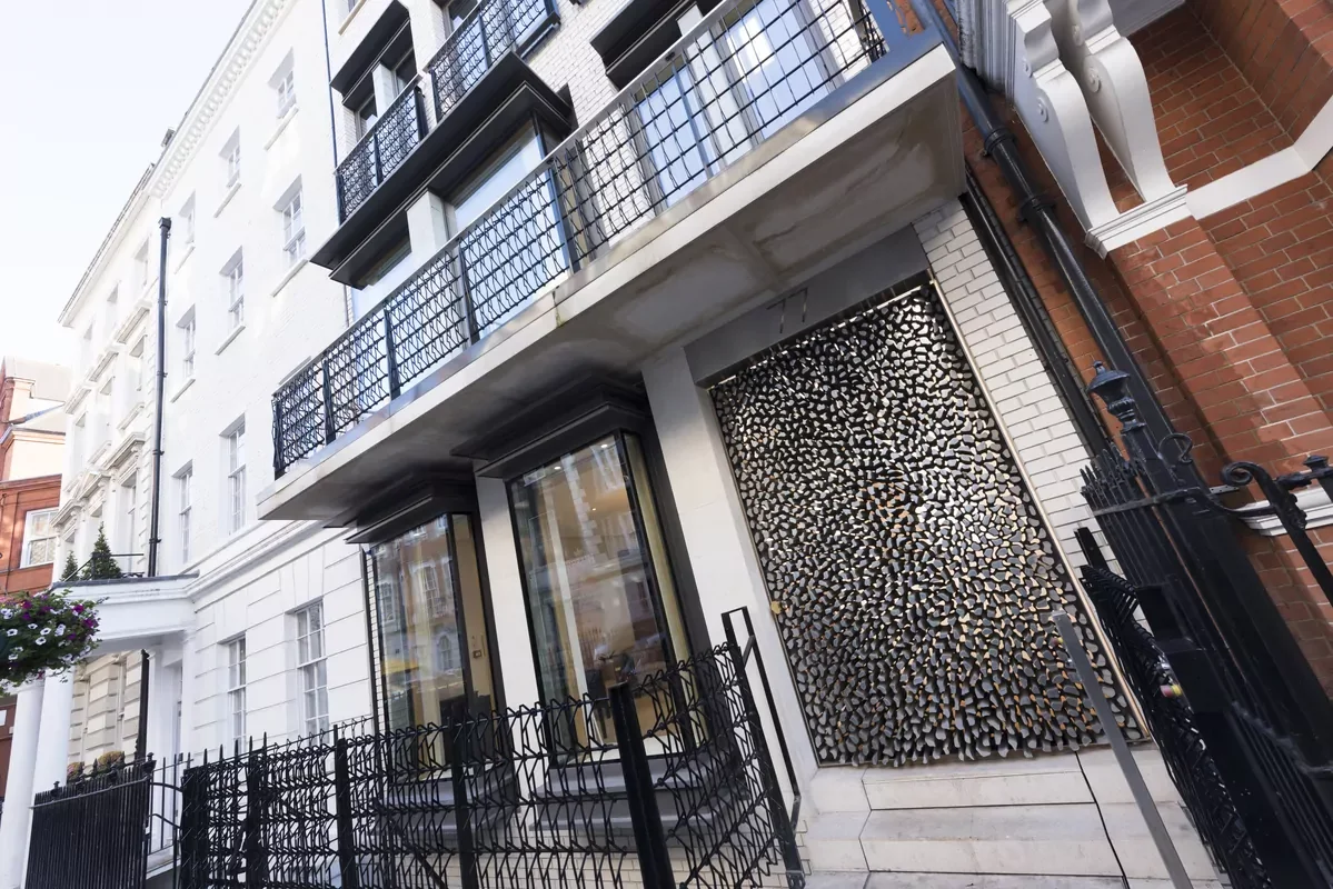 The image shows the front of a modern multi-story building with a decorative metal screen on the ground floor, glass doors, and multiple balconies with black railings. There are neighboring buildings and a street visible.