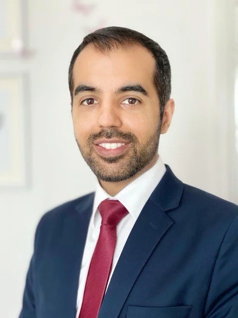 Professional headshot of a man with dark hair, beard, wearing a navy suit, white shirt, and red tie, smiling in an office setting.