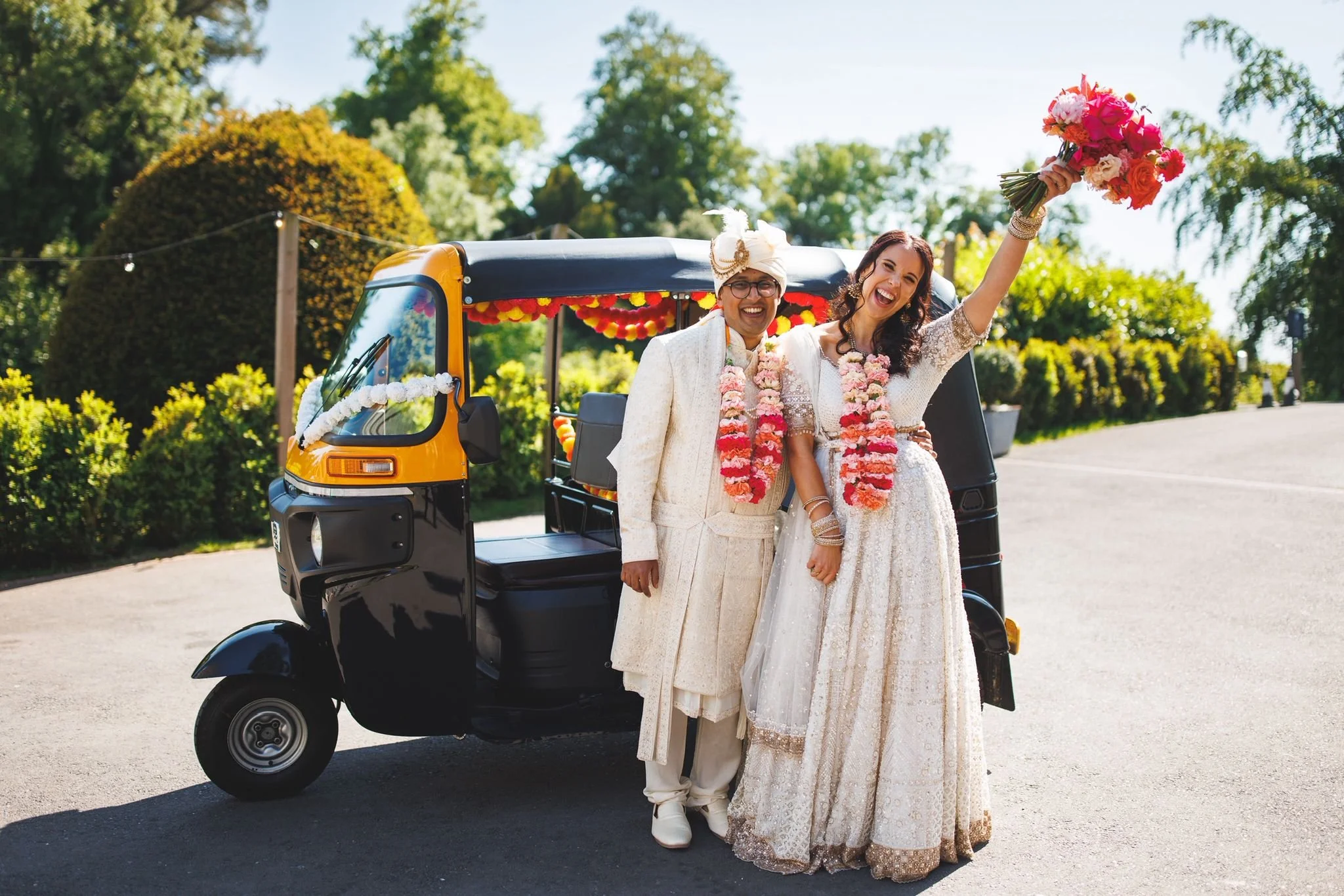 A couple celebrating wedding in traditional Indian attire standing next to an auto rickshaw decorated with flowers during daytime in a lush green outdoor setting.