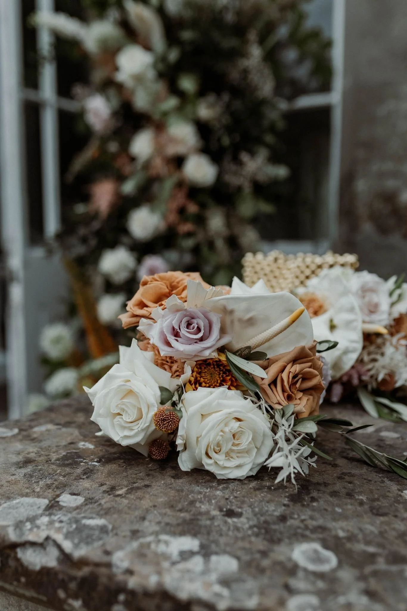A bouquet of white, peach, and blush pink flowers, including roses and other blooms, placed on a rustic stone surface with a floral arrangement in the background.
