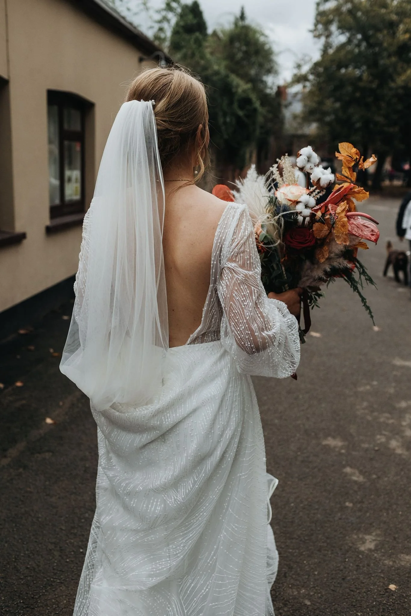 A bride in a white, backless wedding dress holding a bouquet of flowers, standing outdoors on a paved street with trees and buildings in the background.