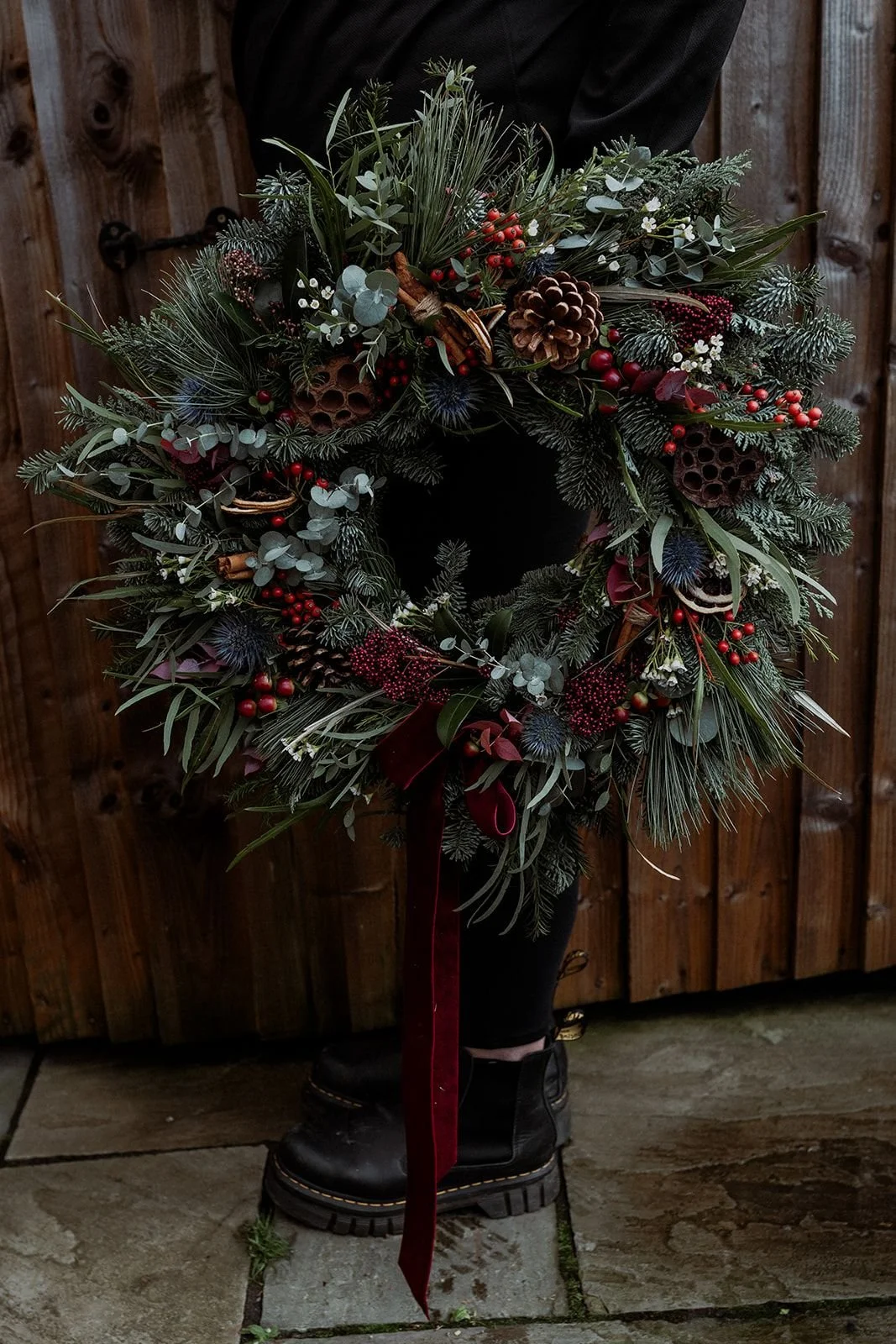 A person is holding a Christmas wreath filled with evergreen branches, red berries, pinecones, cinnamon sticks, and other greenery, with a red ribbon hanging down, in front of a wooden fence.