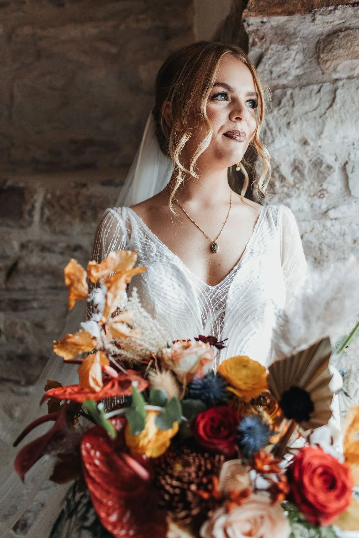 A woman in a wedding dress holding a colorful bouquet of flowers, standing by a stone wall.