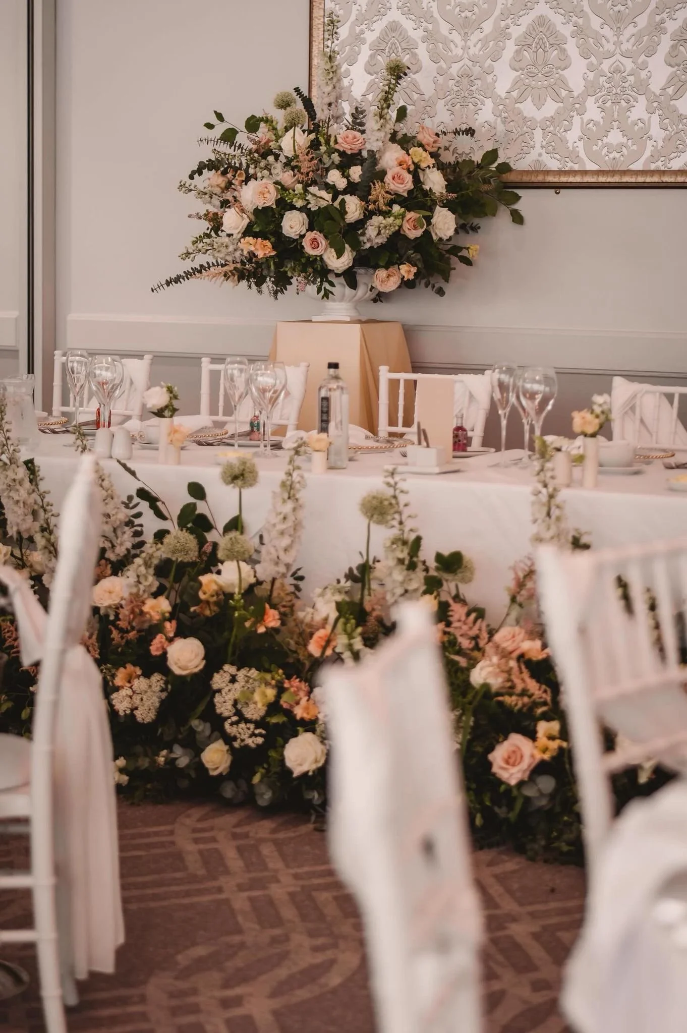 A decorated banquet table with floral arrangements, glassware, and place settings, set for a formal event or wedding reception, with a large floral display on a stand behind the table.
