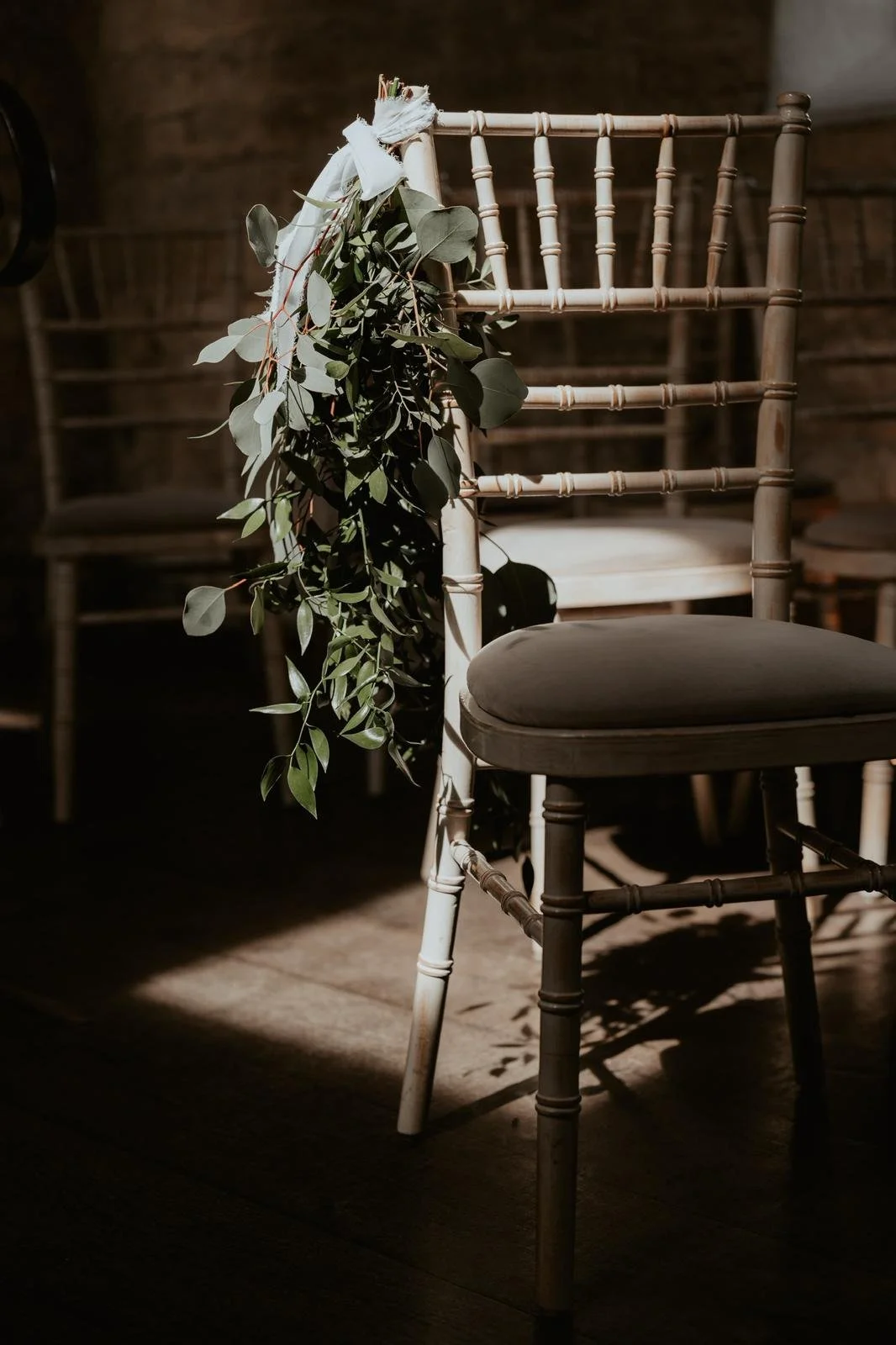 A white standing chair with a dark cushioned seat decorated with green leafy foliage tied to the backrest, in a dimly lit room with warm lighting and wooden flooring.