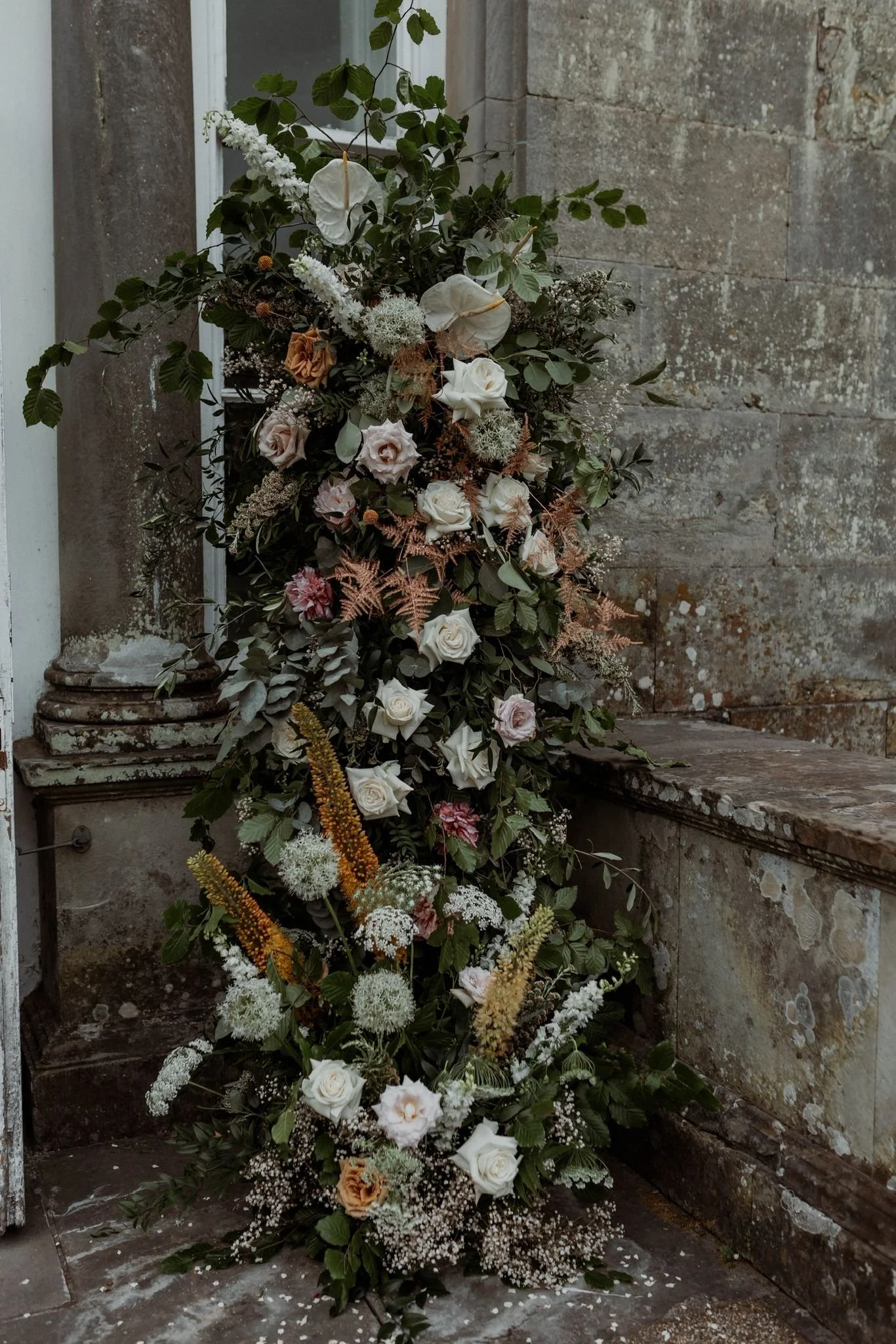 A vertical arrangement of white, pink, and beige flowers with green and brown foliage, placed against an old stone wall and steps.