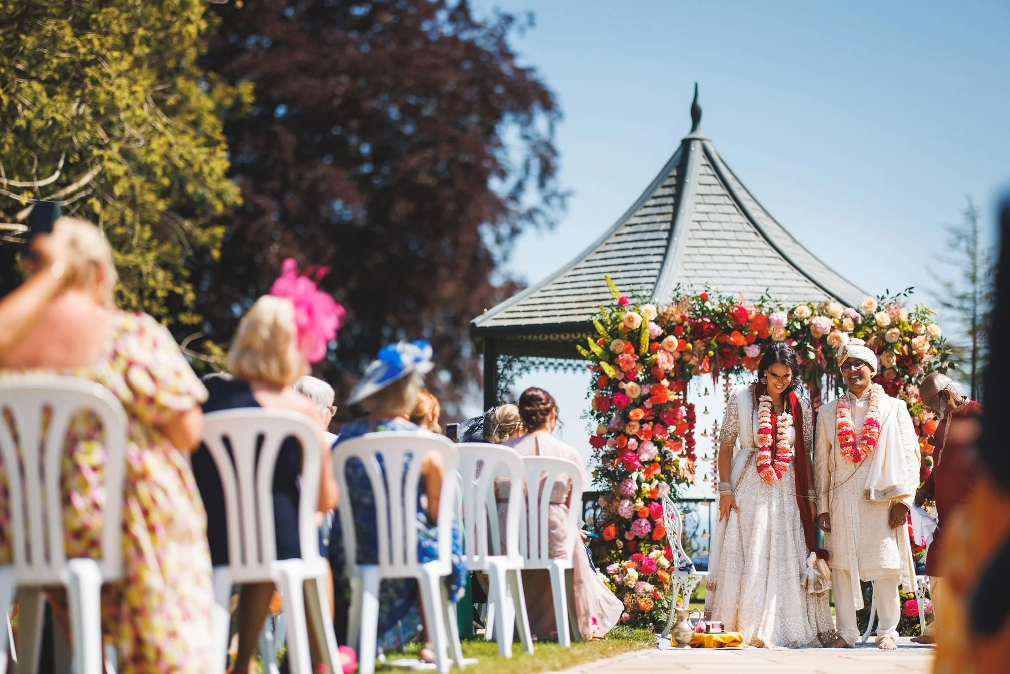 A wedding ceremony with a couple in traditional attire standing in front of a floral arch, with guests seated on white chairs under a gazebo on a sunny day.