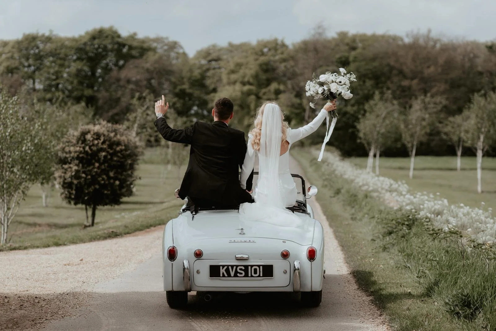 A bride and groom riding in a vintage white convertible car on a country road, with the bride holding a bouquet of white flowers and both waving.