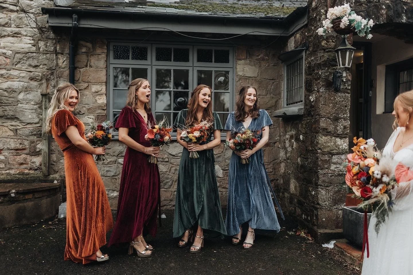 Four women in colorful dresses holding bouquets of flowers are smiling and laughing outside in front of a stone building, as one woman in a white dress holds a bouquet and interacts with them.