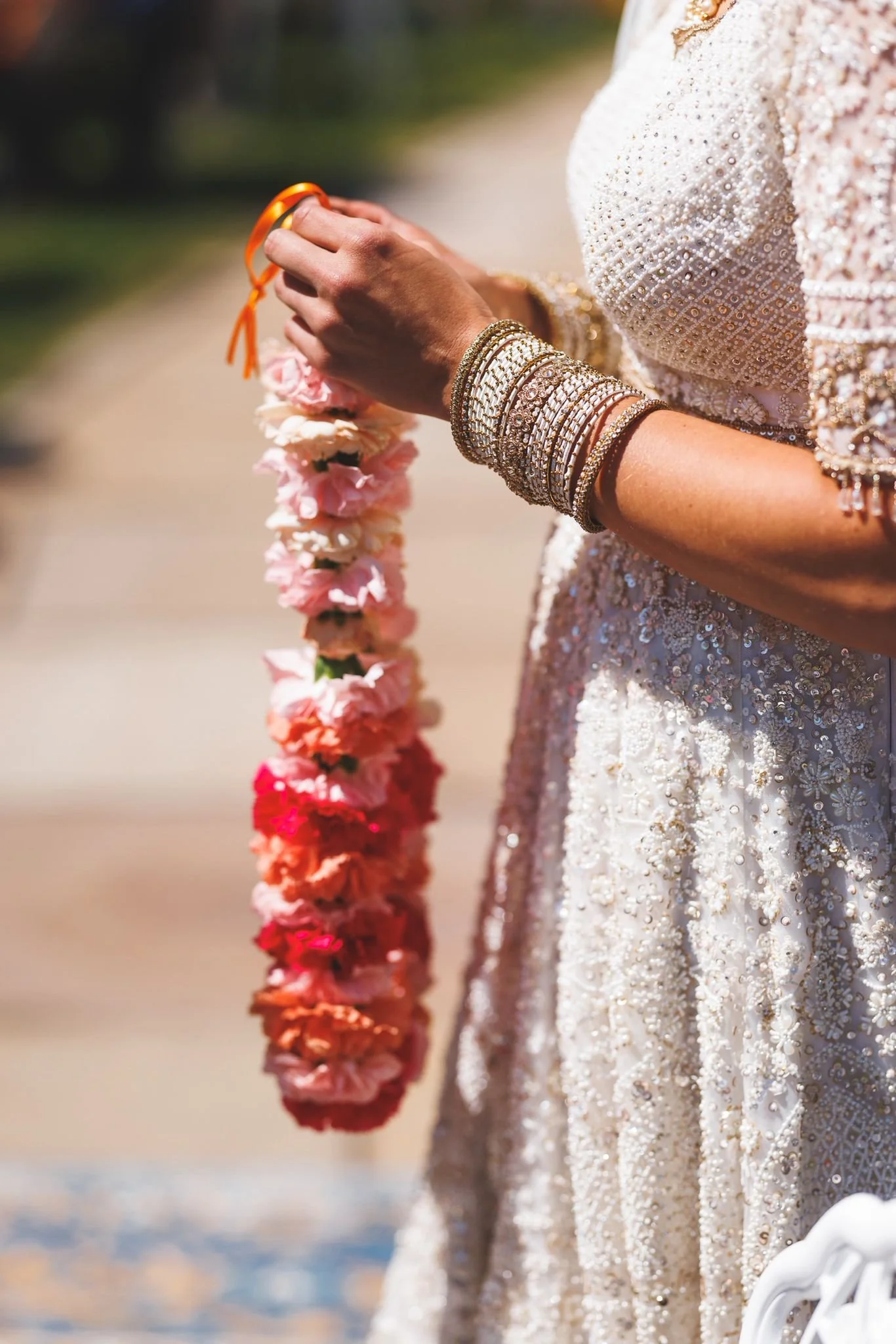 A woman in an ornate, beaded dress holds a pink and orange flower garland with both hands, wearing multiple bracelets on her right wrist.