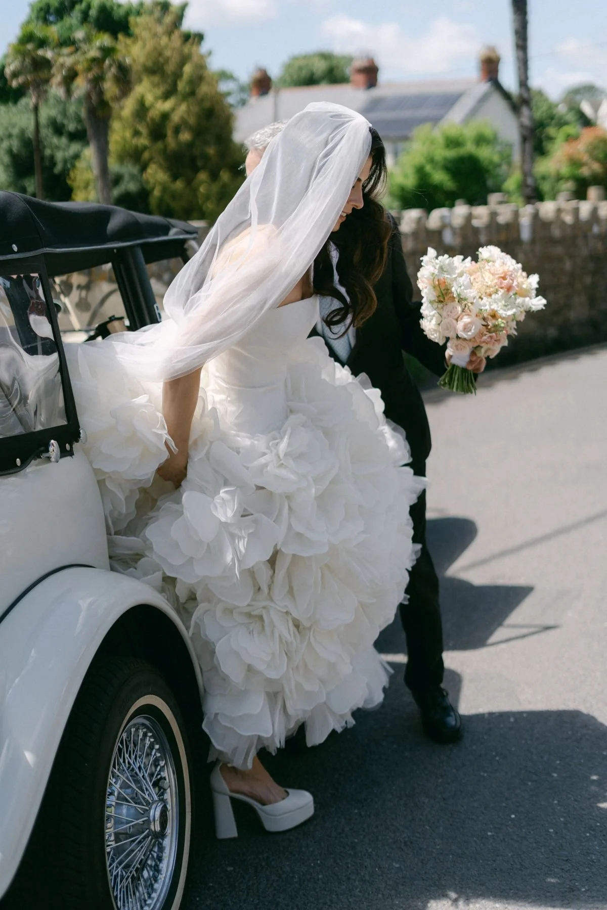 Bride in a white wedding gown and veil stepping out of a vintage car with a bouquet of flowers, accompanied by a person in a black suit, on a sunny day with trees and houses in the background.