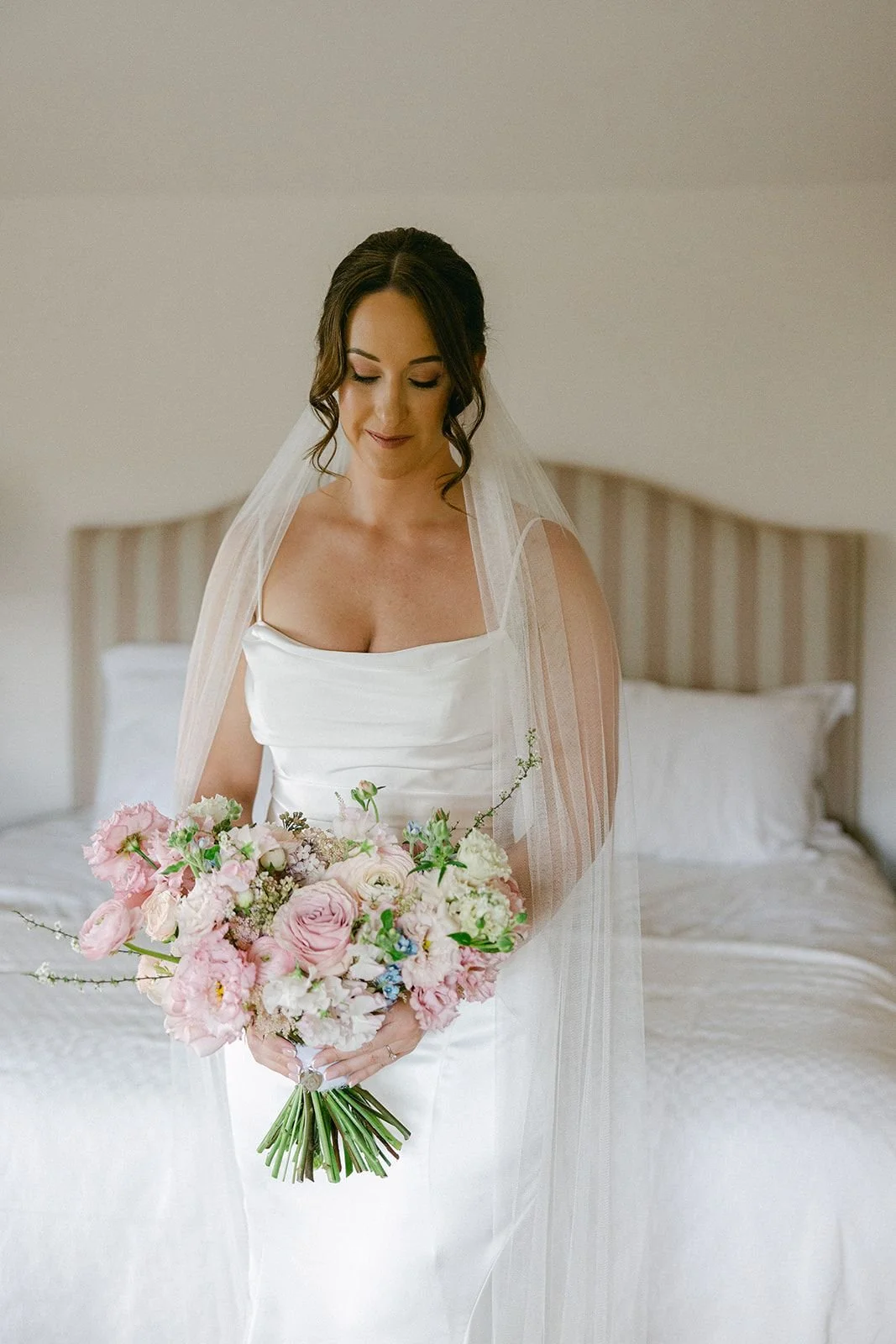 A bride in a white wedding gown holding a bouquet of pink and white flowers, with a wedding veil, standing in a softly lit bedroom.