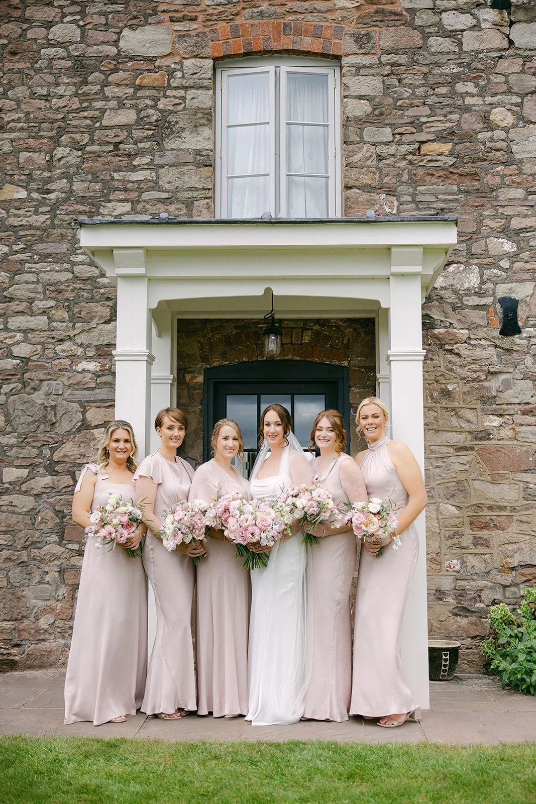 A bride with five bridesmaids standing in front of a stone house with a black door, holding pink and white bouquets, smiling for a wedding photo.