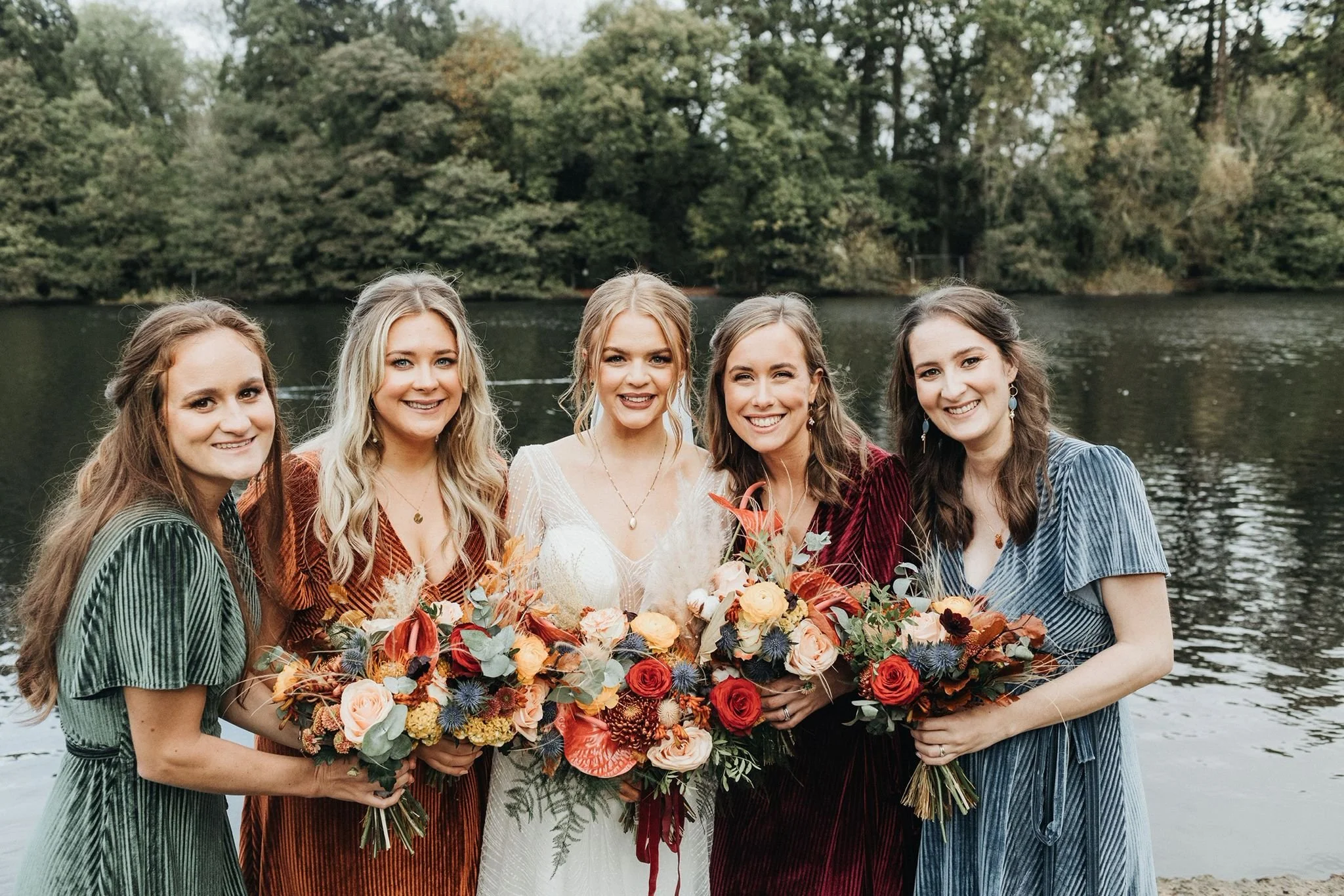 Five women in dresses holding bouquets of flowers stand by a lake with trees in the background.