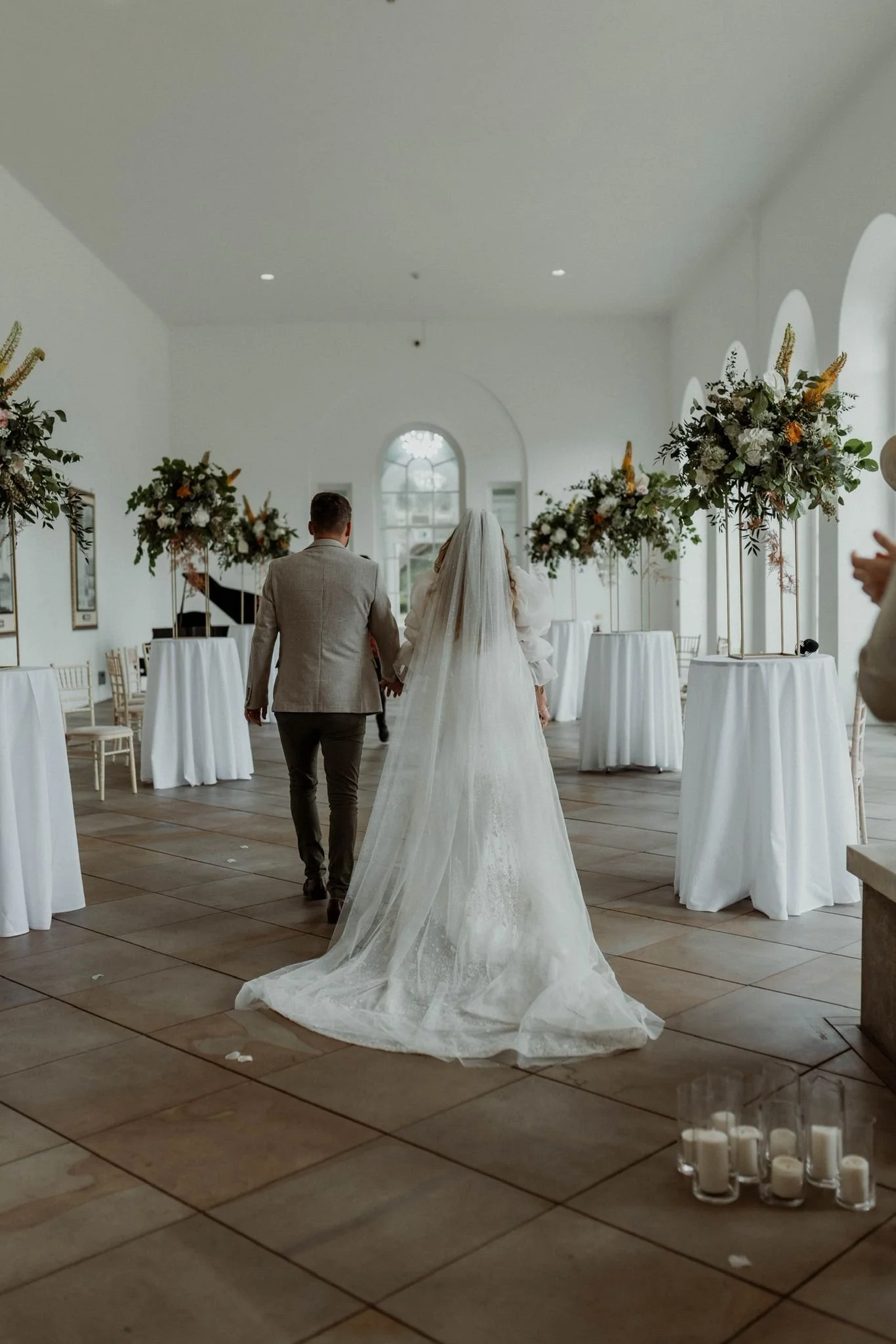 A bride and groom walking hand in hand in a decorated wedding venue with white walls, arched windows, and tall floral arrangements on round tables.