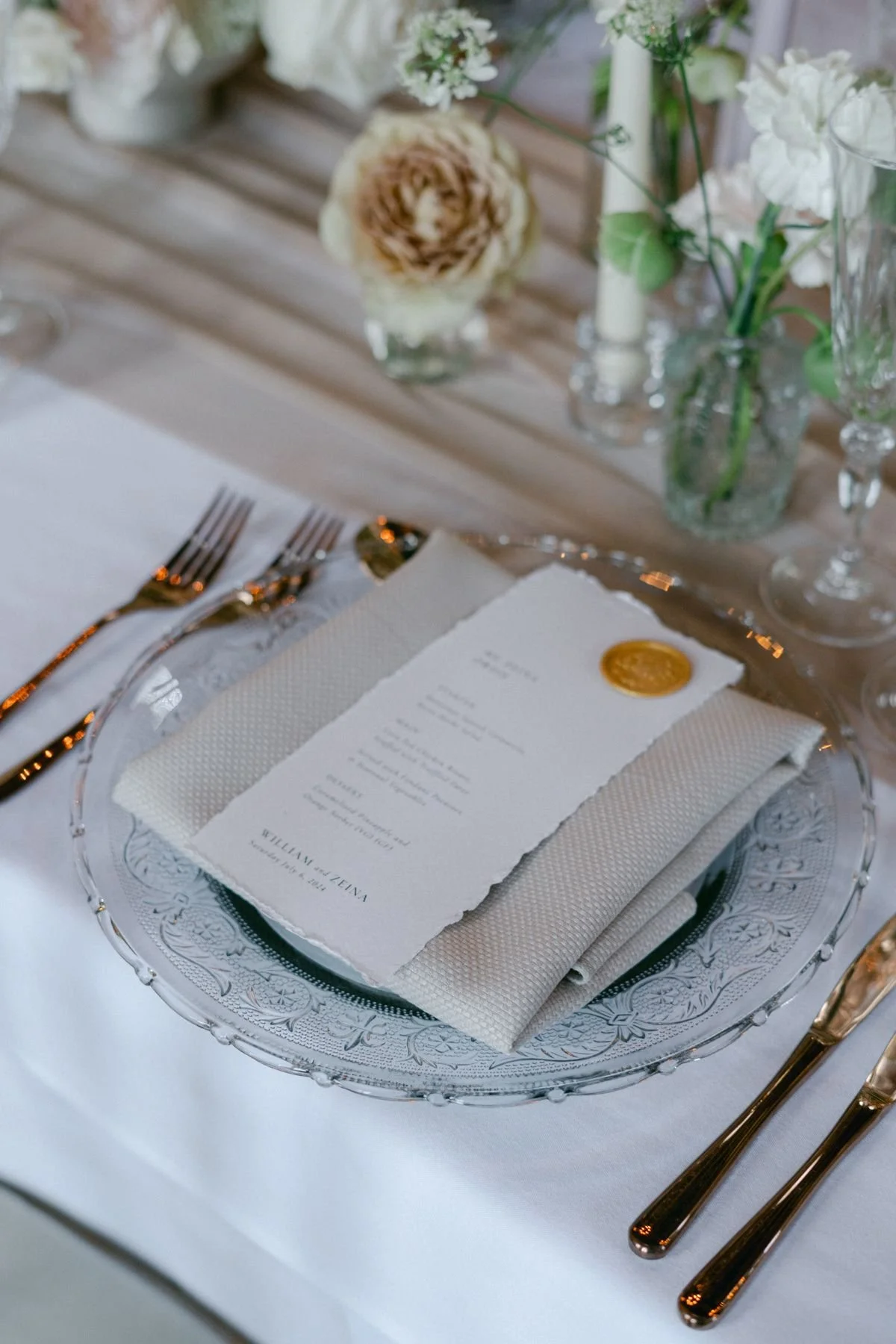 Elegant wedding table setting with a menu on a white plate, gold cutlery, and floral arrangements in the background.