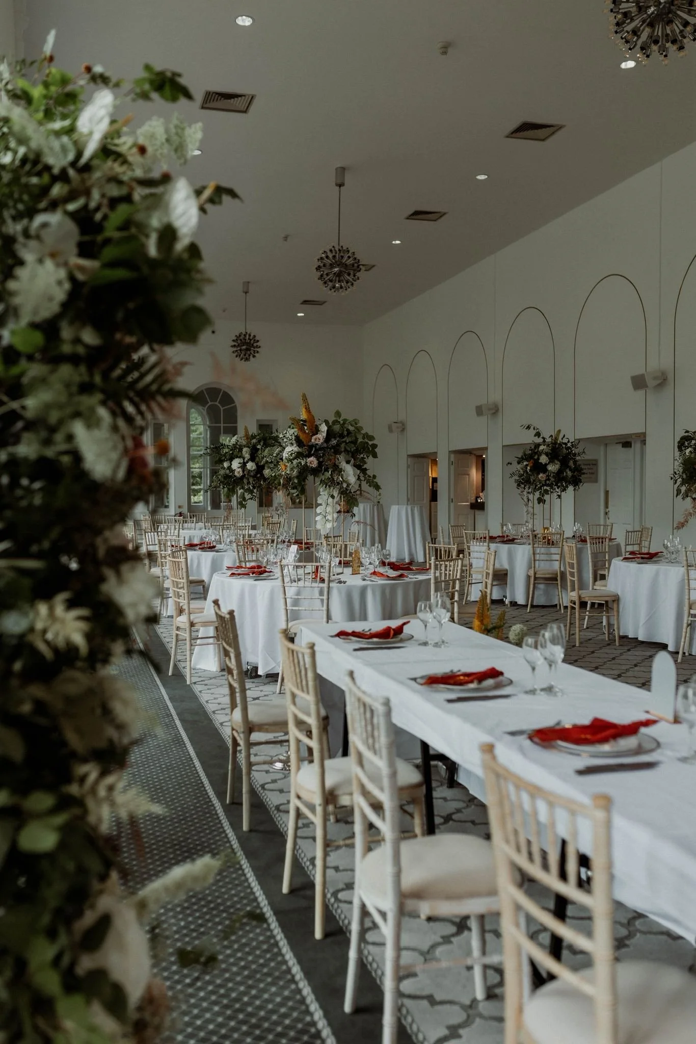 Elegant banquet hall decorated with tall floral centerpieces, set for a formal event with white tablecloths, gold chairs, red napkins, wine glasses, and plates.