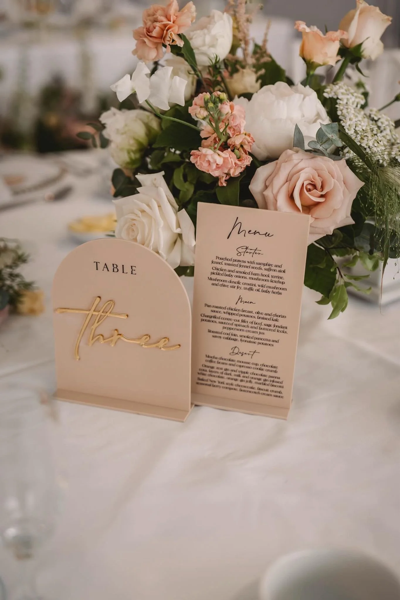 Elegant table setting featuring a floral centerpiece with roses and greenery, a menu card, and a table number sign that reads 'three' in gold script.