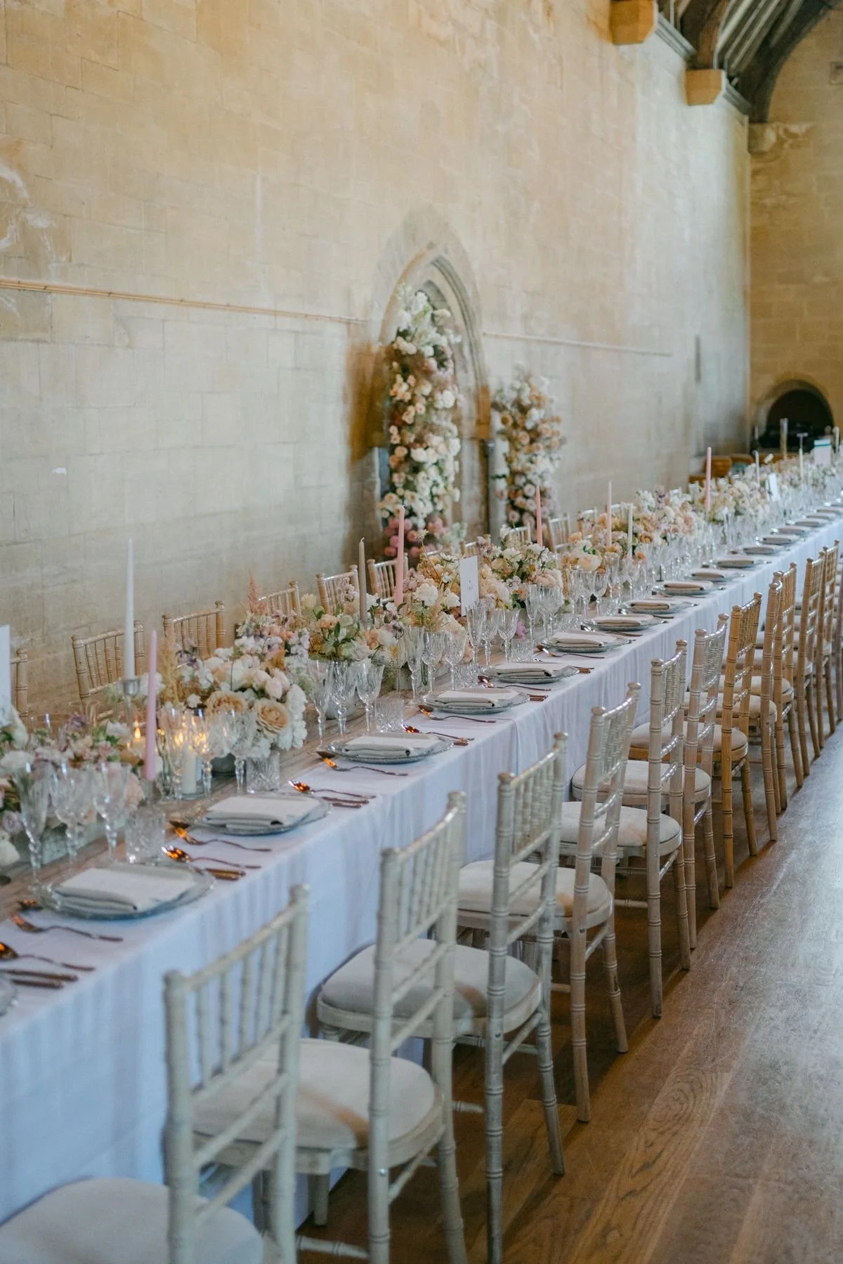 Elegant wedding reception table set with white tablecloth, floral centerpieces, candleholders, and glassware, against a stone wall with floral arrangements in background.