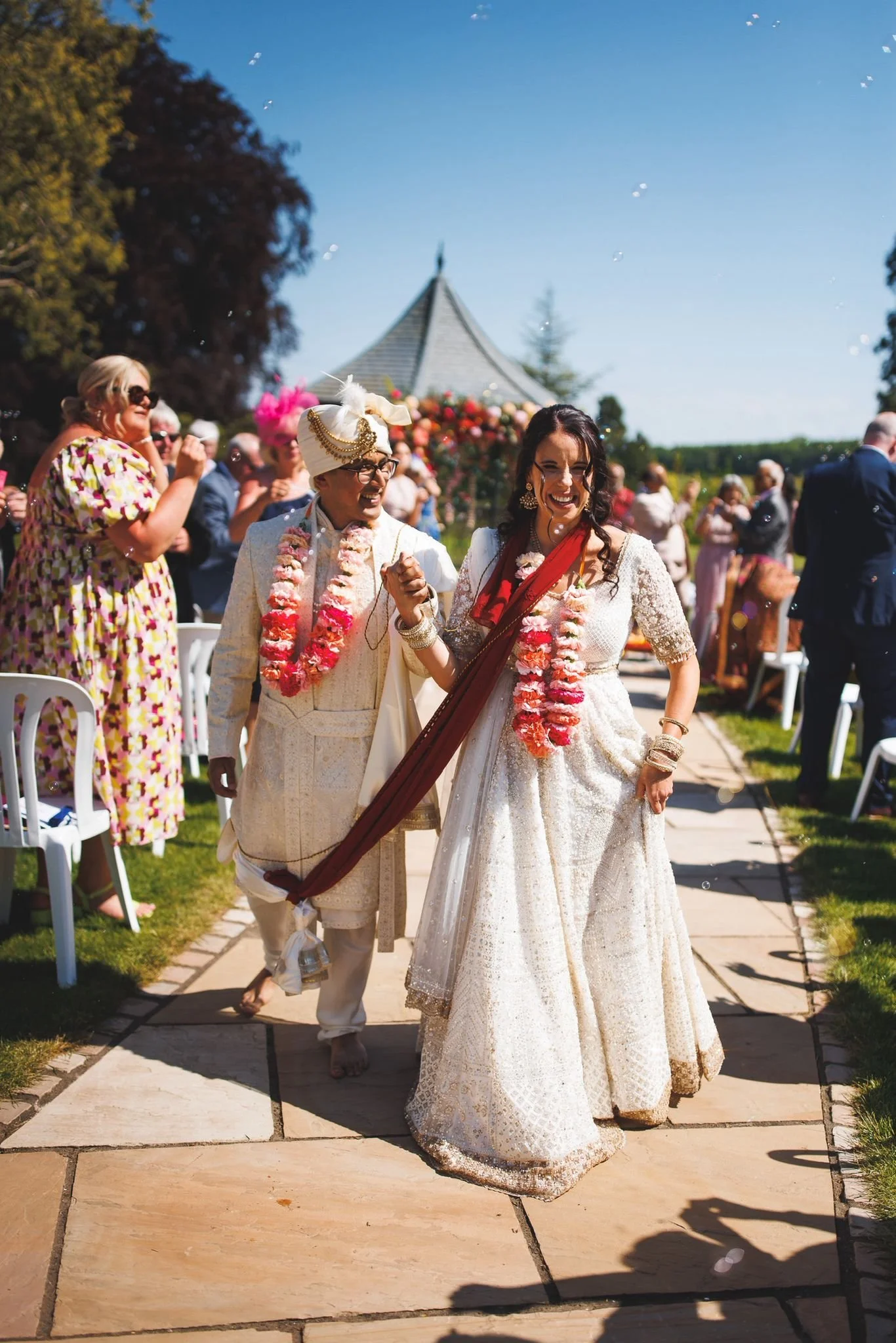Bride and groom walking down the aisle at an outdoor wedding ceremony, smiling and holding hands, with guests celebrating around them and a small structure and trees in the background.