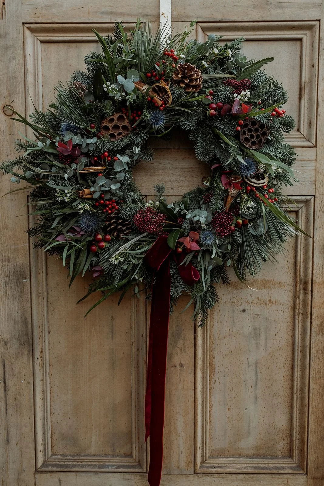 A festive Christmas wreath made of greenery, pinecones, red berries, and dried seed pods, hanging on a wooden door with a red ribbon bow at the bottom.