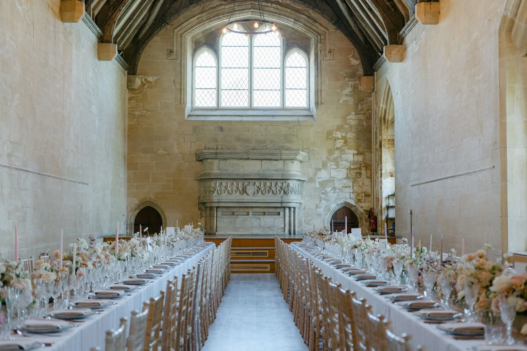 Long banquet tables decorated with pink and white floral arrangements and tableware inside a historic stone building with a large window and stone fireplace.