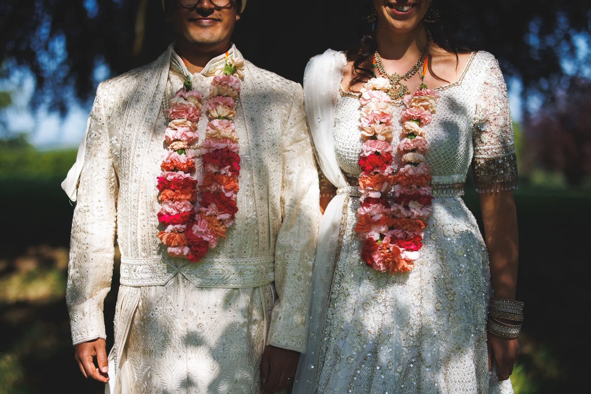 A bride and groom in traditional wedding attire with floral garlands, standing outdoors on a sunny day.
