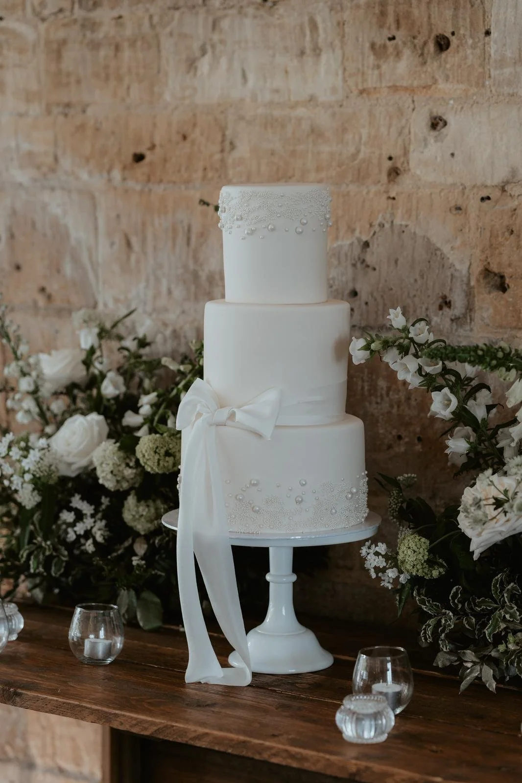 A white three-tiered wedding cake with decorative pearls and a bow, placed on a white cake stand in front of a brick wall, surrounded by white floral arrangements and small candles.