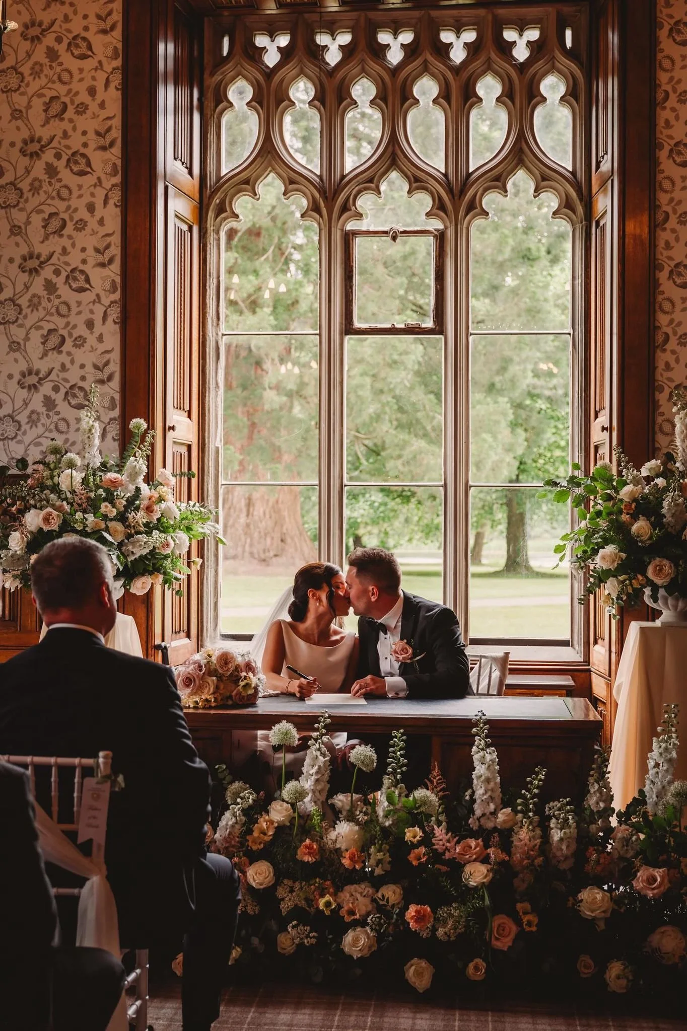 A bride and groom sharing a kiss during their wedding ceremony, seated at a table in front of large ornate windows with a scenic outdoor view, surrounded by floral arrangements.