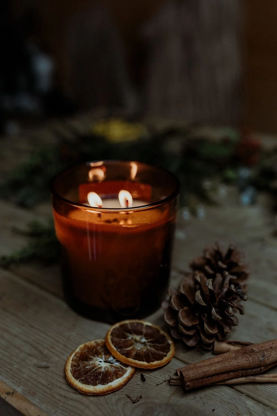 A lit candle in a brown glass holder surrounded by dried orange slices, a pine cone, and cinnamon sticks on a wooden surface, with blurred greenery in the background.