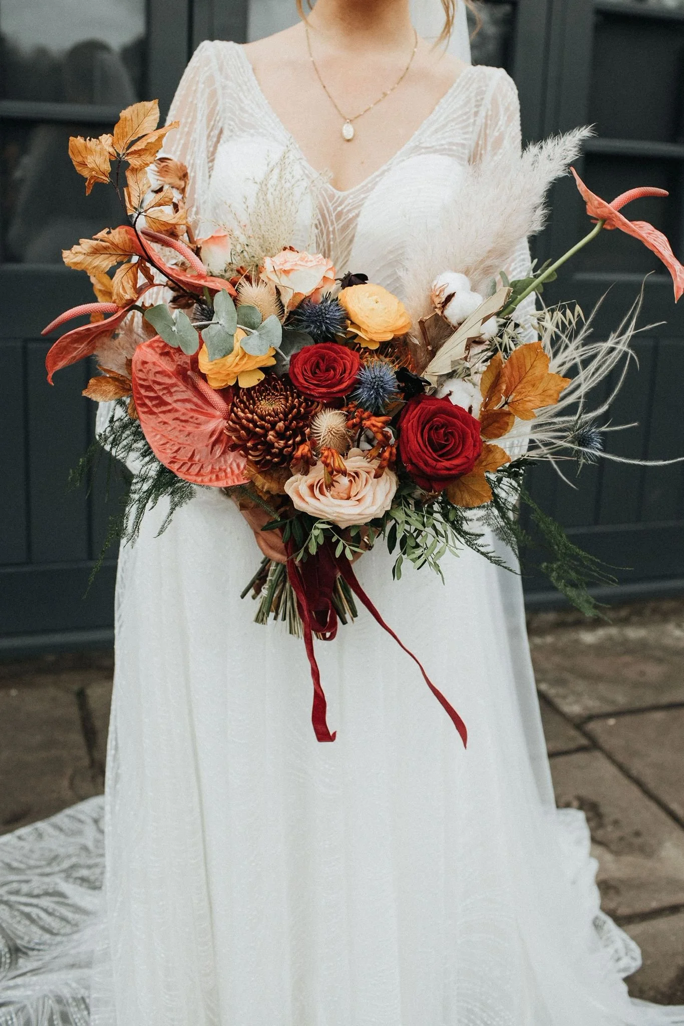 A bride holding a large bouquet of mixed flowers in front of her, wearing a white wedding dress and a gold necklace with a pearl pendant.