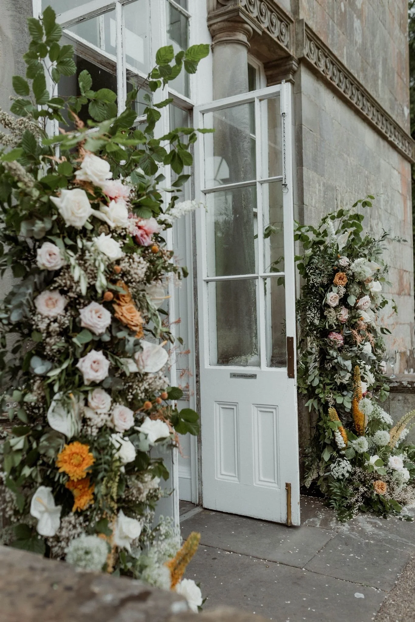 Open white door with glass panes, flanked by tall floral arrangements with white, pink, orange, and yellow flowers, set against a stone building.