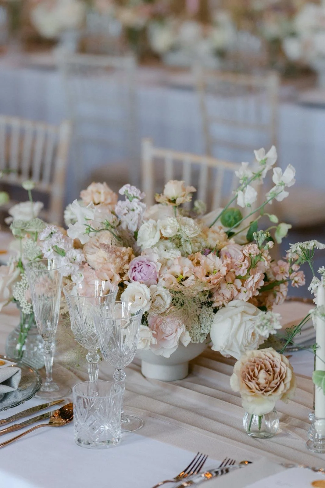 Elegant table centerpiece with a large bouquet of pastel-colored flowers in a white vase, surrounded by crystal glassware and table setting on a light-colored tablecloth at a formal event.